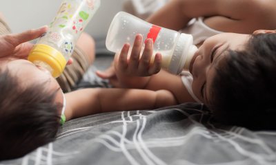 Two babies lying on their backs, each drinking formula from bottles