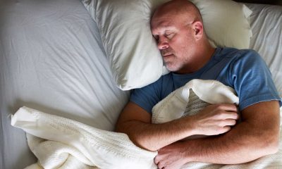 White man with shaved head and gray stubble wearing a blue T-shirt asleep in bed,
