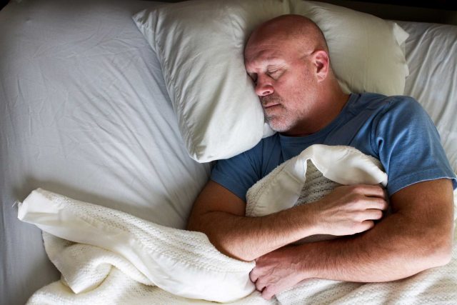 White man with shaved head and gray stubble wearing a blue T-shirt asleep in bed,
