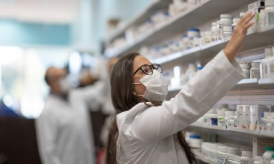 A Pharmacist with a mask on reaches for a bottle of medicine on a shelf above her head