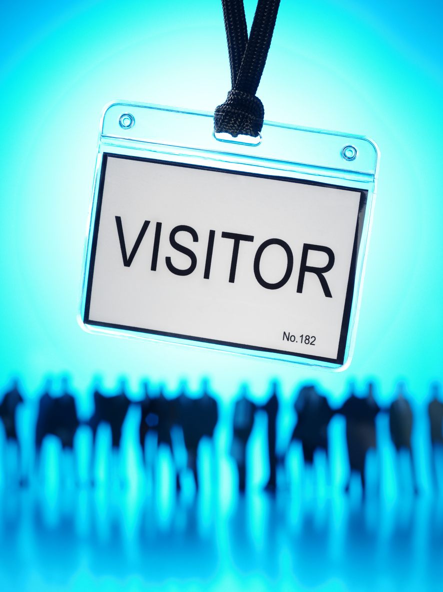A badge attached to a lanyard with the word "Visitor" in the foreground, as a group of silhouetted people stand in line in the background.