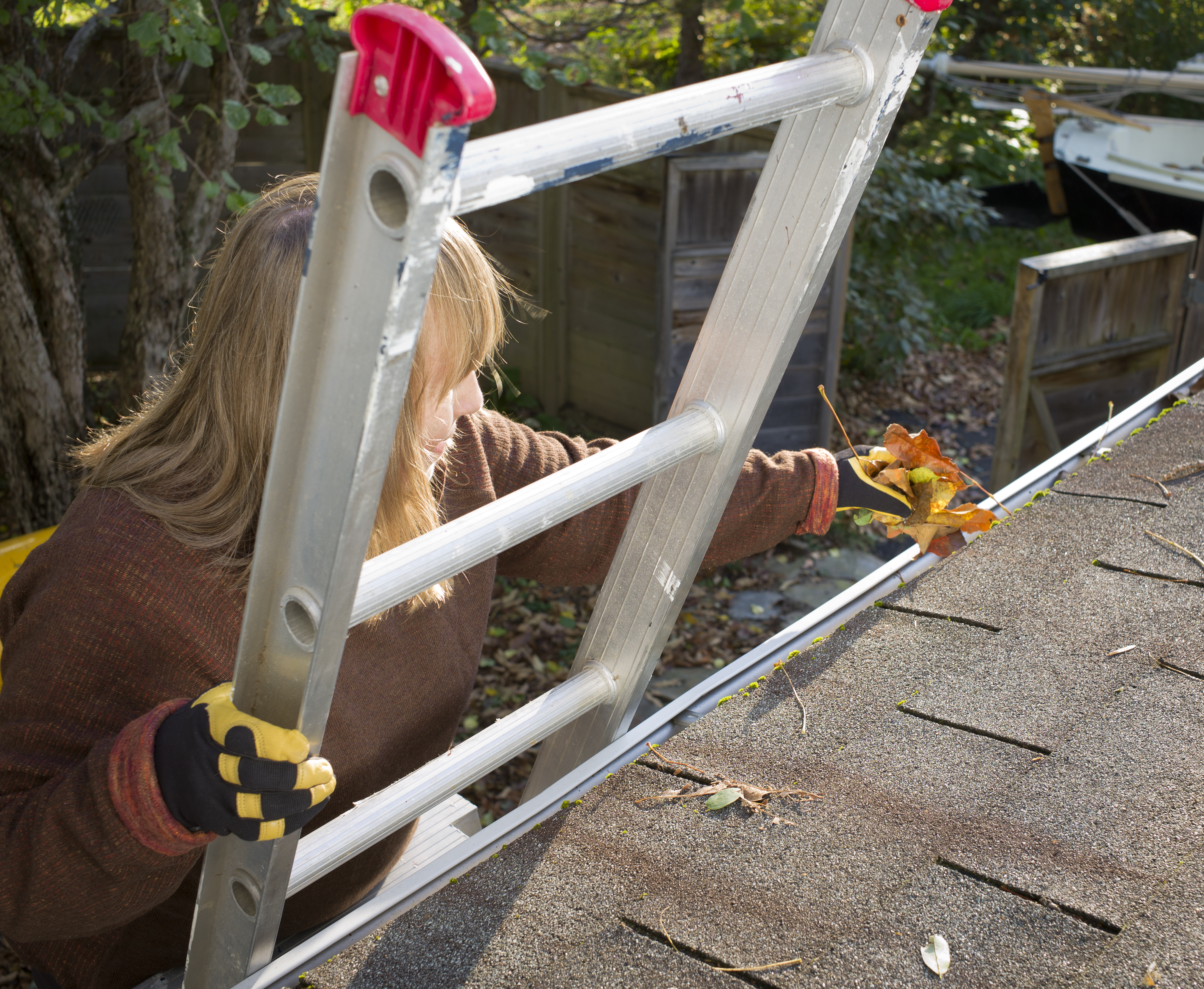 Woman cleaning leaves out of gutter on a ladder.