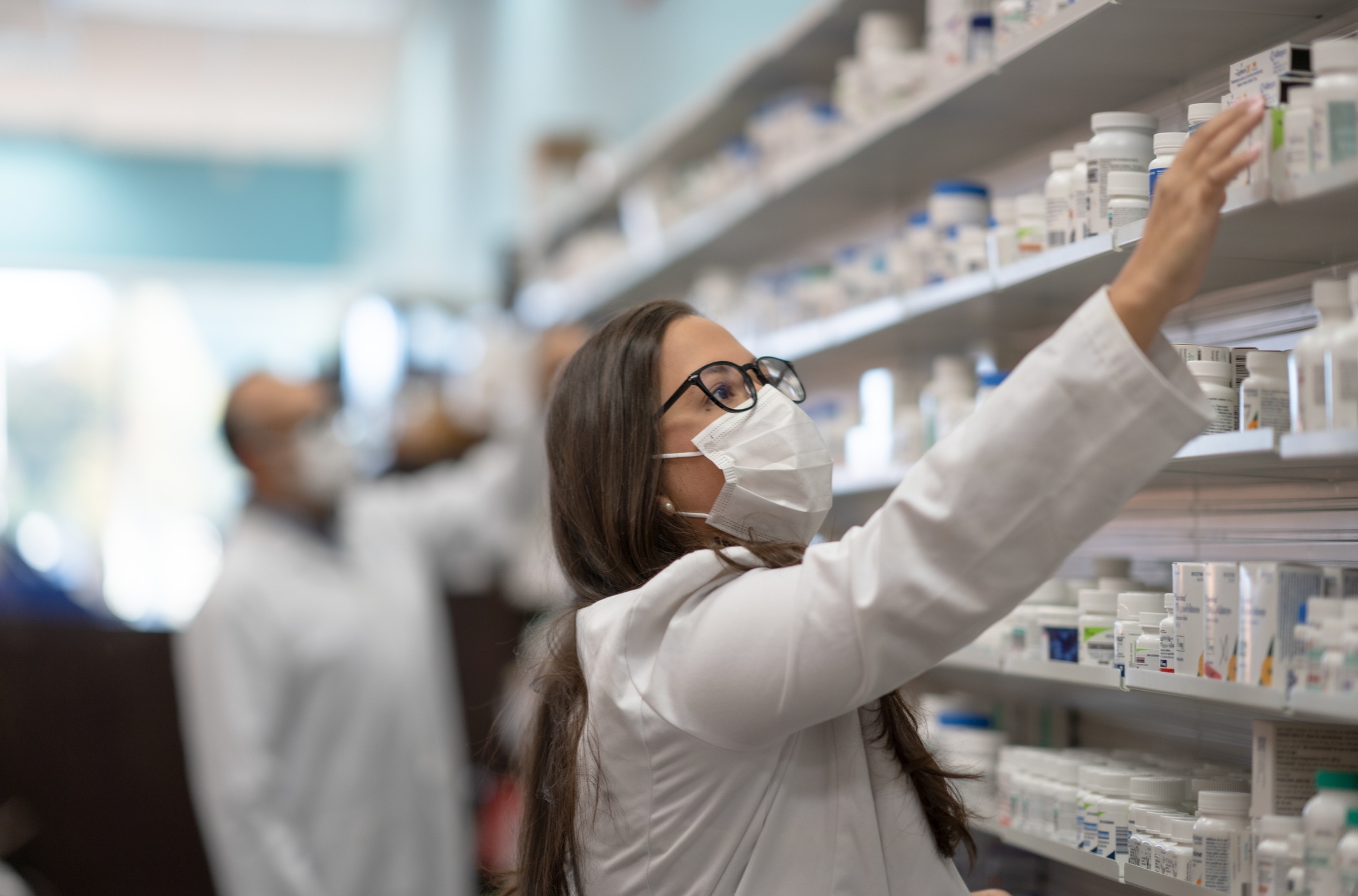 A Pharmacist with a mask on reaches for a bottle of medicine on a shelf above her head