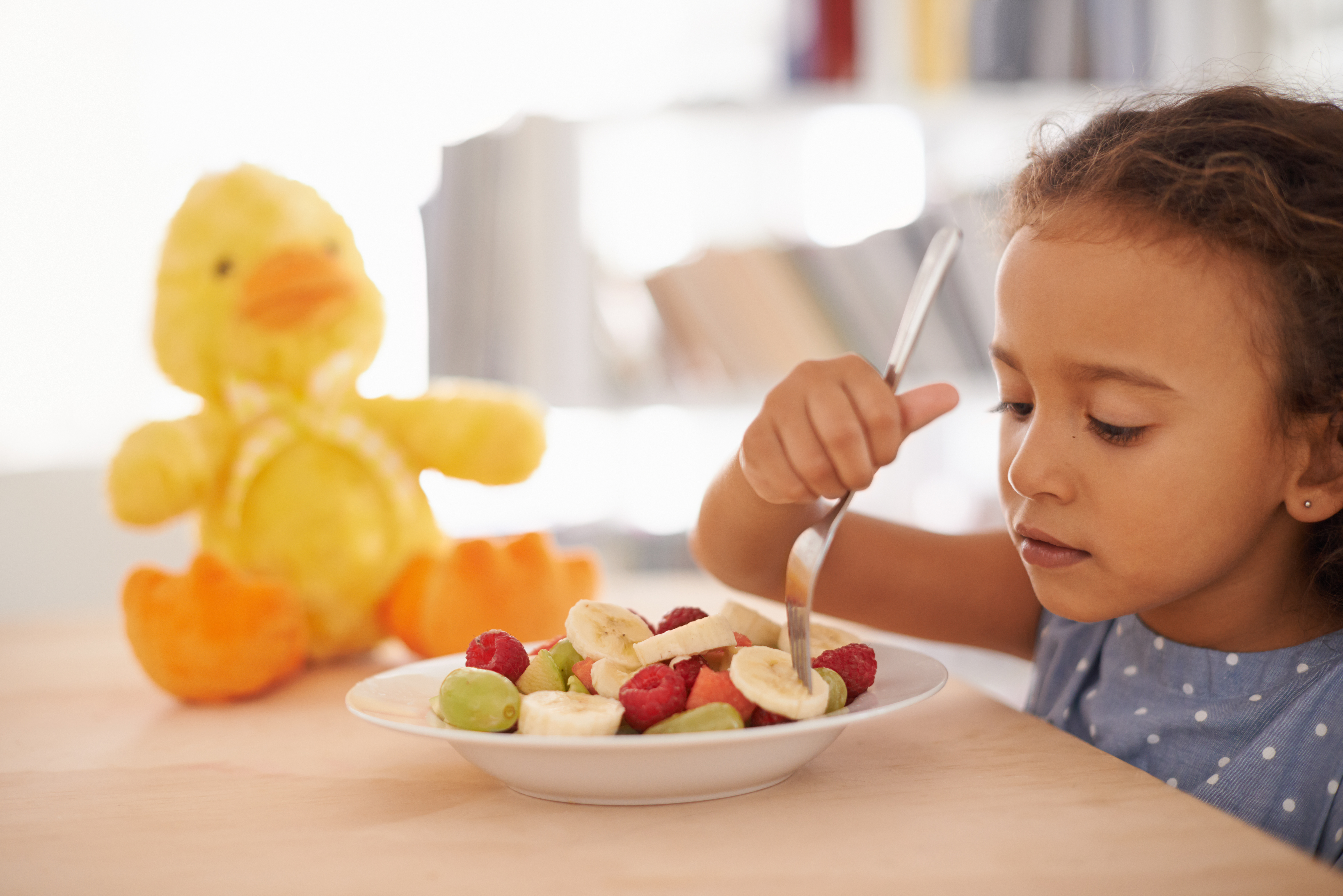 A child picking through fruit on a plate with their fork as a rubber duck sits in the background