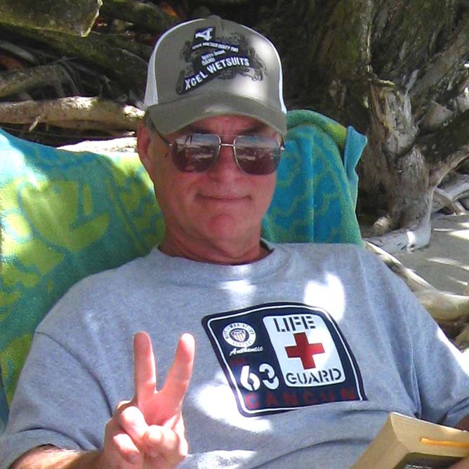 A grinning man relaxing in a chair with a hat and sunglasses displays the peace sign