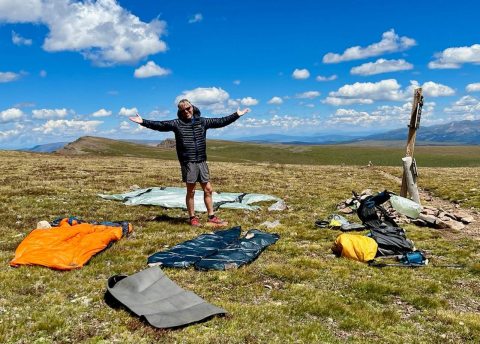 Adult man stands amid several sleeping bags outdoors in remote area.