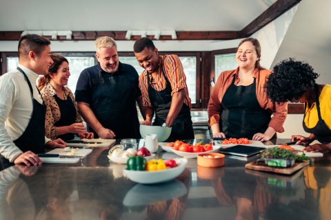 A group of jovial people gathered around a table in a kitchen, prepping food to make a meal