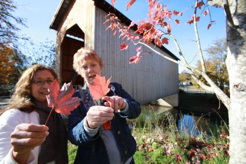 Two women hold up colorful red autumn leaves beside a wooden covered bridge on a sunny fall day.