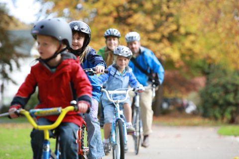 Family of five wearing bicycle helmets rides together along a paved path in Eugene, Oregon, surrounded by colorful autumn trees.