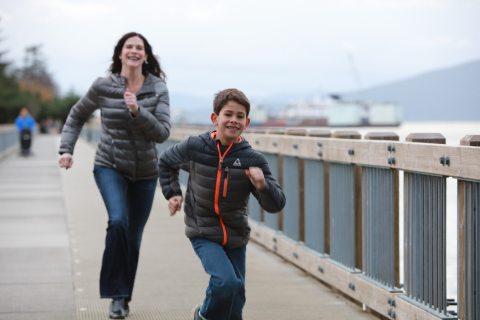 Smiling young boy runs along a waterfront boardwalk with a woman running behind him, both wearing jackets on an overcast day in Bellingham, Washington