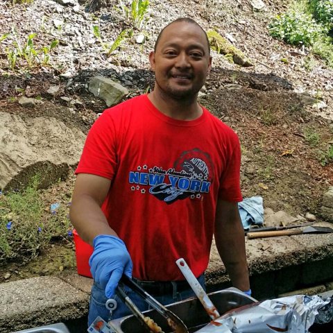 A man in a red shirt, serving dinner outside, stops to smile for a photo