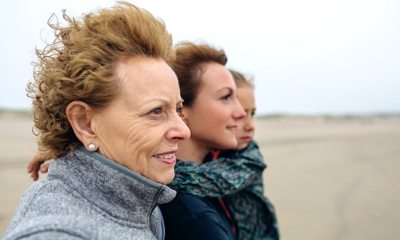 Three generations of women stand on a beach looking into the distance