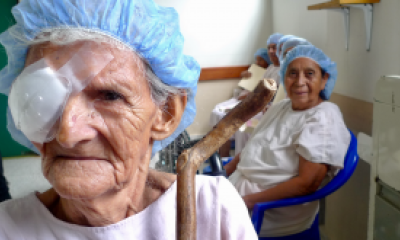 An woman in the background smiles and looks on as an older woman in the foreground with a medical eye covering faces the camera
