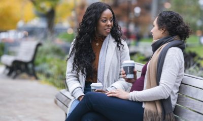 Two women sit on a park bench drinking coffee and having a conversation