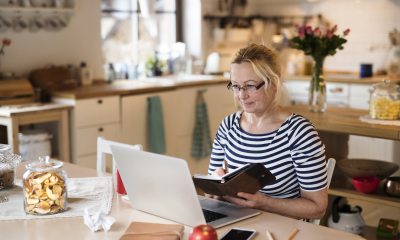 Older woman sits at kitchen table, looking at laptop screen and paper calendar