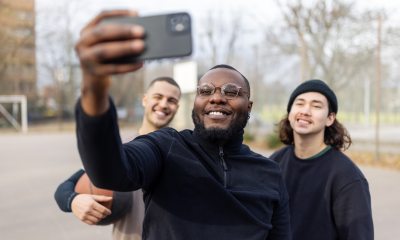 Black man wearing glasses uses cell phone to take selfie with 2 other men on outdoor basketball court