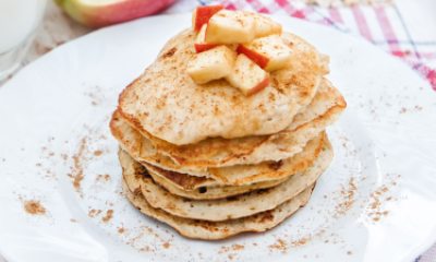 White plate with a stack of apple cinnamon pancakes. There is also a cup of milk and a fork on the table.