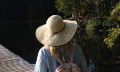 A blonde woman wearing a wide-brim sun hat and a backpack walks away from the camera along a dock at the lake 