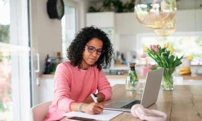 Woman wearing glasses sits at kitchen table, writing with a pen next to laptop