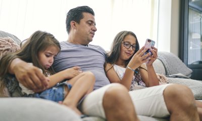 Father sits on couch looking at phone screen with young daughters