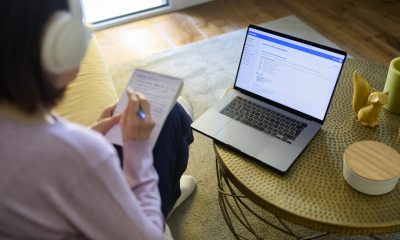 A woman with audio headphones sits in front of a laptop while writing on a notepad