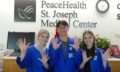 Three PeaceHealth Volunteers standing in front of the St. Joseph Medical Center sign, posing, with their hands up