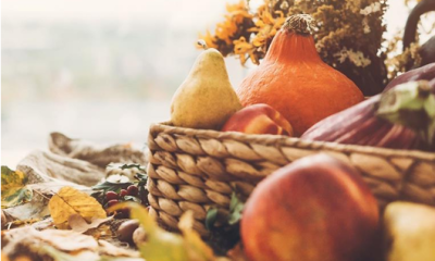 A fall season themed basket with orange, yellow and brown colored vegetables sit next to a pile of leaves