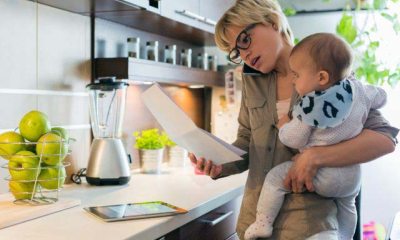 A busy mother on her phone, holds her baby in the kitchen while looking at a piece of paper