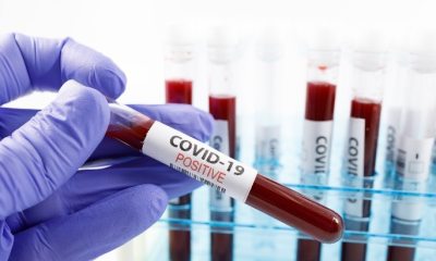 Close-up of lab workers gloved hand holding a filled glass test tube with the words "COVID-19 Positive" visible on the side