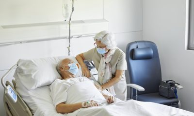 An older patient lies in bed while being comforted by an elderly visitor