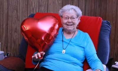 An elderly woman smiles while sitting and holding a heart shaped balloon