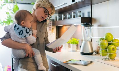 A woman holding a baby while in the kitchen looks at a piece of paper and talks on her mobile phone