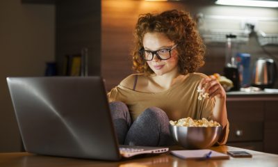 A young adult watches a laptop in a dimly lit kitchen, with popcorn on her left