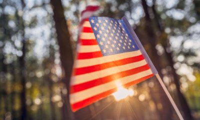A United States flag waves in front of sunlight beaming through trees