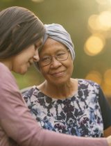 Two women outdoors -- one young and one older -- embrace and rest their for foreheads against each other. The older woman wears a headwrap indicating hair loss from cancer treatment.
