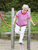 Smiling woman with short white hair balances on a piece of outdoor playground equipment