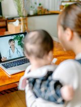 A woman holds her baby while talking to a doctor via a telehealth appointment.