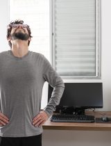 Bearded man in long-sleeve gray shirt looks up to ceiling, stretching back in front of desk