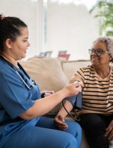Dark-haired healthcare provider in blue scrubs takes blood pressure reading of older Black patient sitting on couch