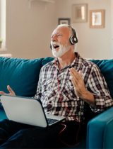 A man sitting on a couch with a laptop and headphones, singing or reacting jubilantly to the sound