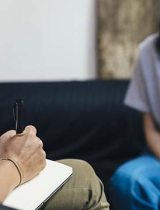 A person takes notes while another in the background speaks