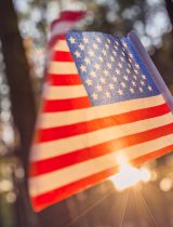 A United States flag waves in front of sunlight beaming through trees