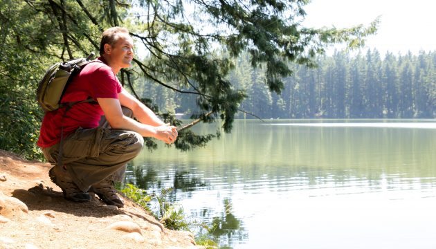 A man in hiking clothing sits on the lake's edge, smiling and looking out at the lake with satisfaction.