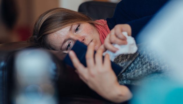 A woman with an illness lying down, looking at her phone in one hand with a tissue in the other hand.