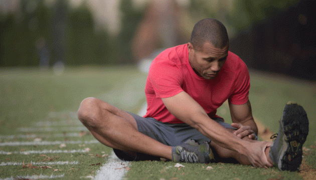 A man stretches his left leg muscle while sitting on a football field sideline.