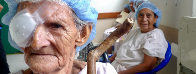An elderly patient in the foreground with a medical eye patch faces the camera as another patient smiles in the background
