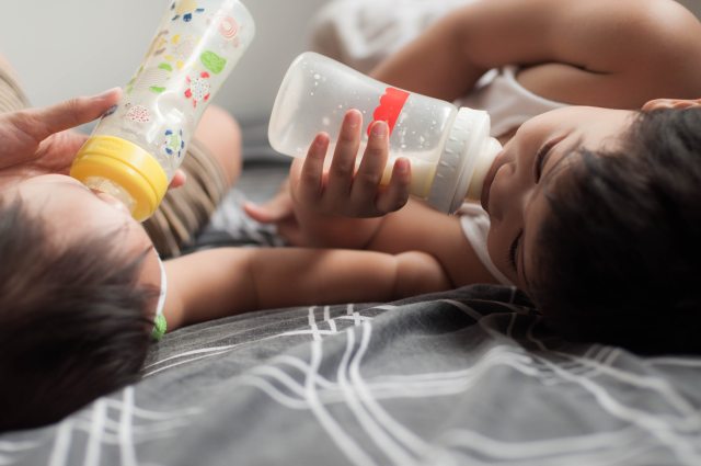 Two babies lying on their backs, each drinking formula from bottles