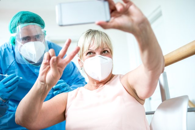 Woman holds up two fingers in a v as she receives vaccination and takes selfie