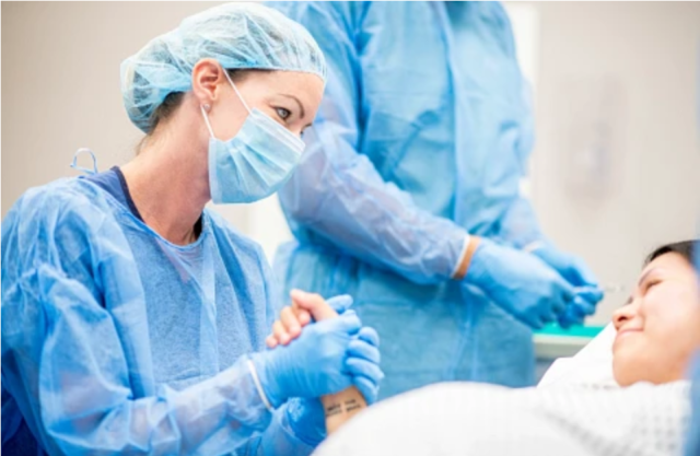 A nurse wearing surgical scrubs holds the hand of a pregnant woman in the OR