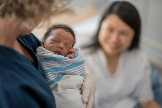 Photo of nurse holding a newborn while smiling mother looks on.